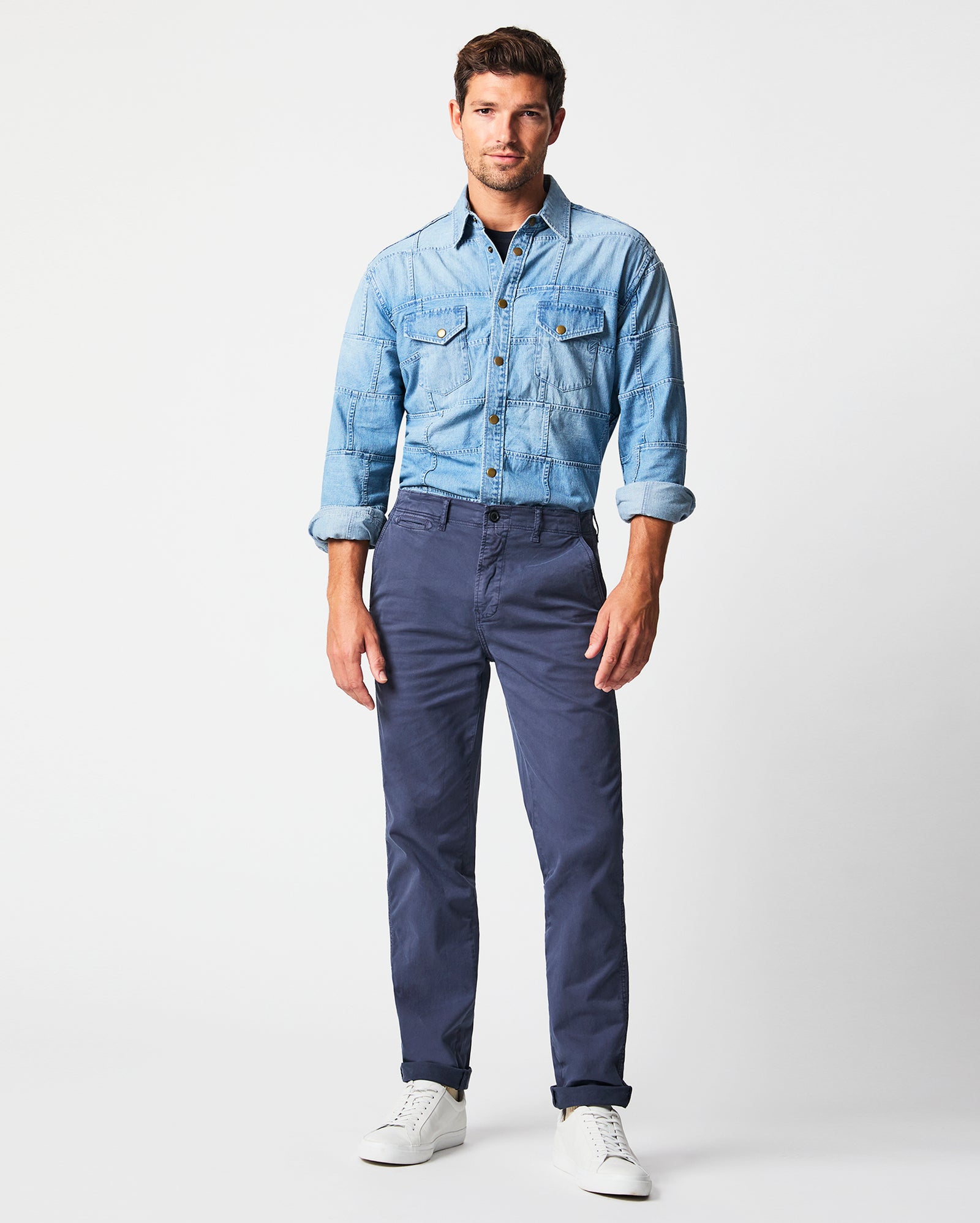 A man stands against a plain white background wearing a light blue denim shirt with rolled-up sleeves, navy blue pants, and white sneakers. He faces the camera with a relaxed expression and hands by his sides.