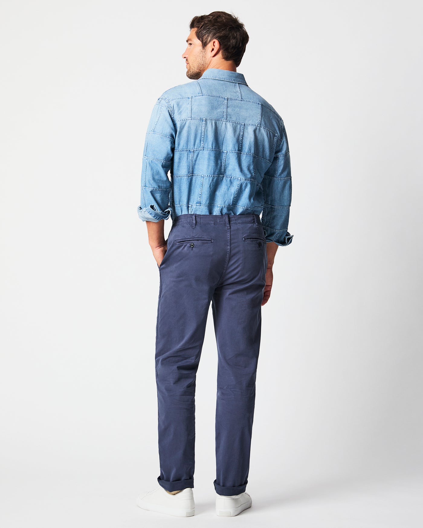 A man stands facing away, wearing a light blue denim shirt with rolled sleeves, tucked into navy chinos, and white sneakers, against a plain white background.