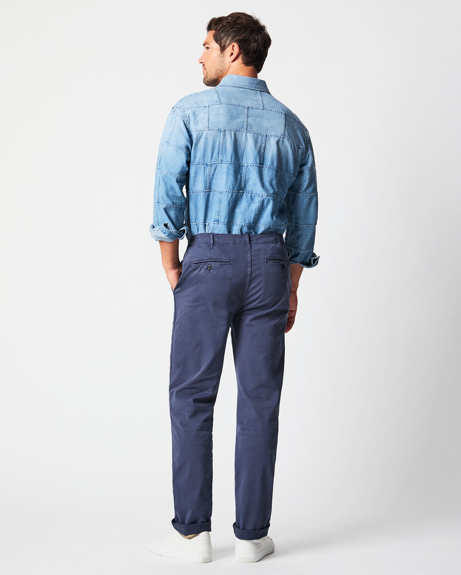 A man stands facing away, wearing a light blue denim shirt with rolled sleeves, tucked into navy chinos, and white sneakers, against a plain white background.