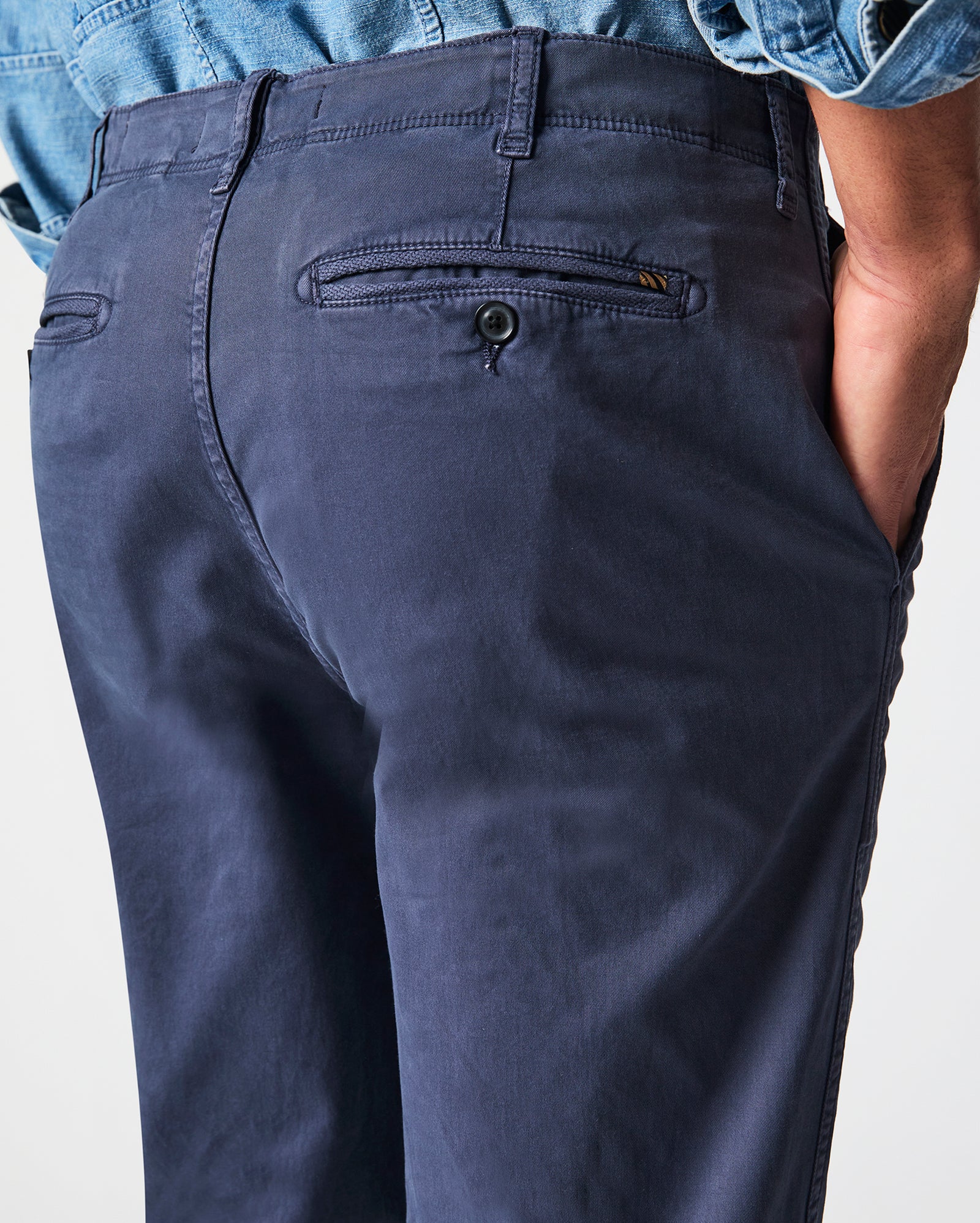 A person wearing navy blue pants and a light blue denim shirt stands with one hand in their pocket, showing the back and side of the pants with buttoned back pockets.