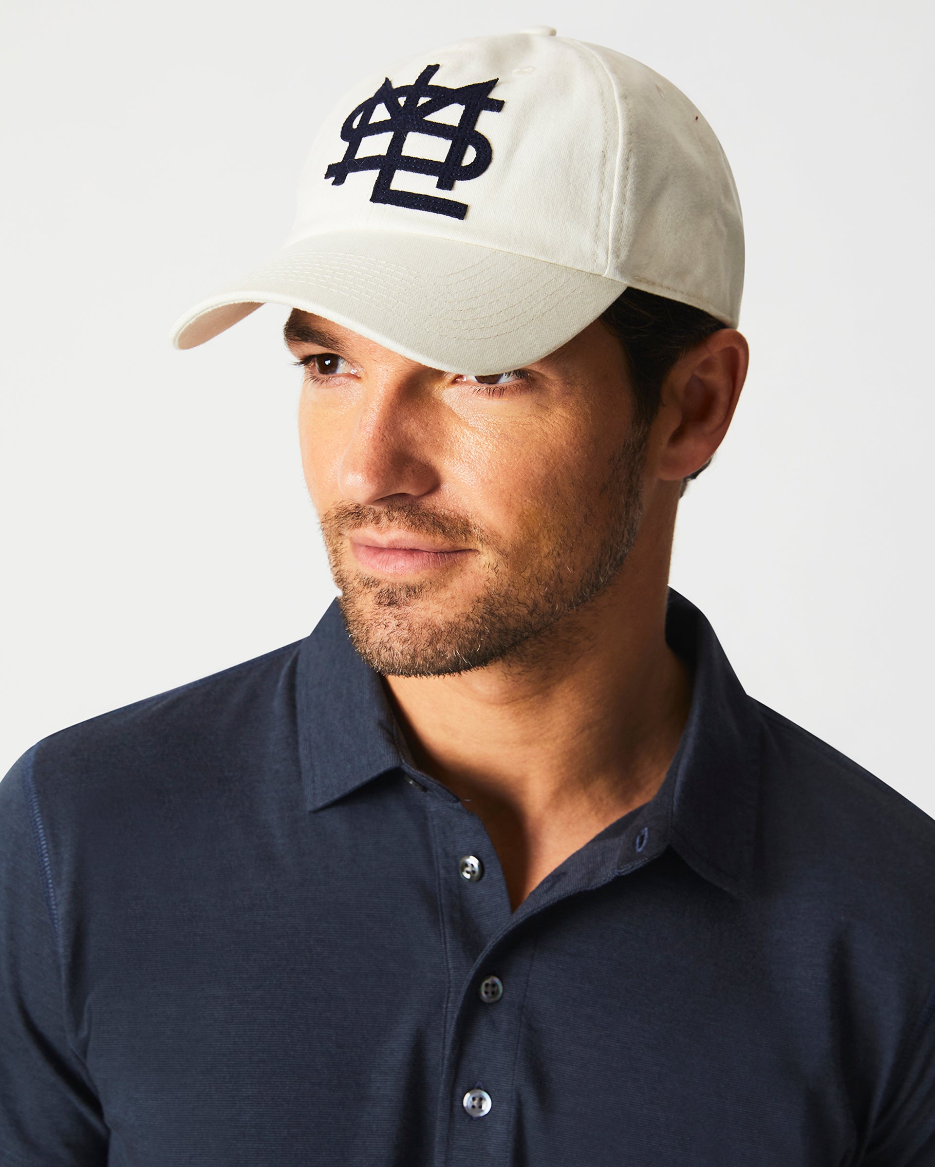A man wearing a cream-colored baseball cap with a navy blue embroidered logo and a navy blue collared shirt looks slightly to the side against a plain background.