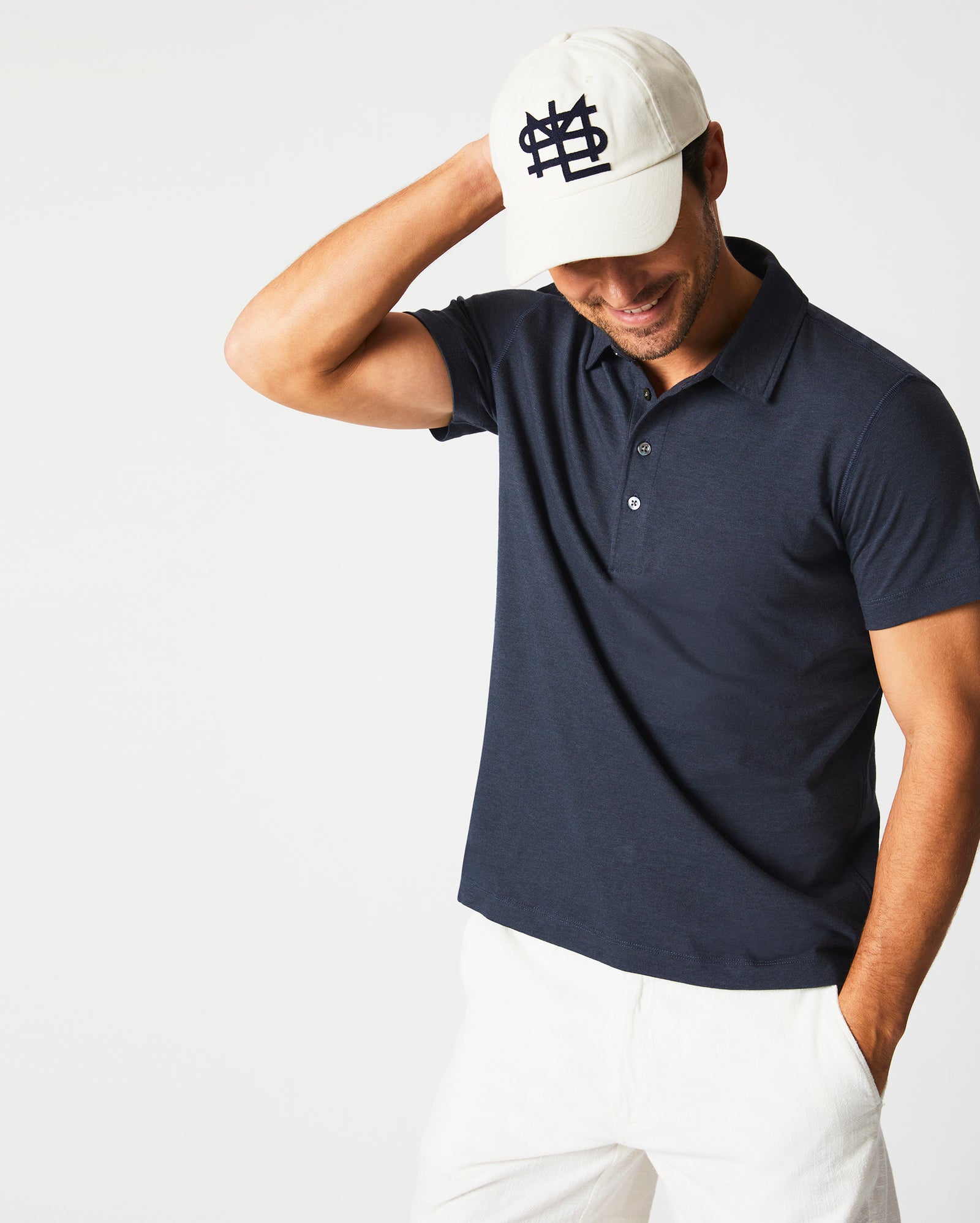 A smiling man in a navy polo shirt and white pants touches the brim of his white baseball cap, which has a dark logo on the front, while standing against a plain white background.