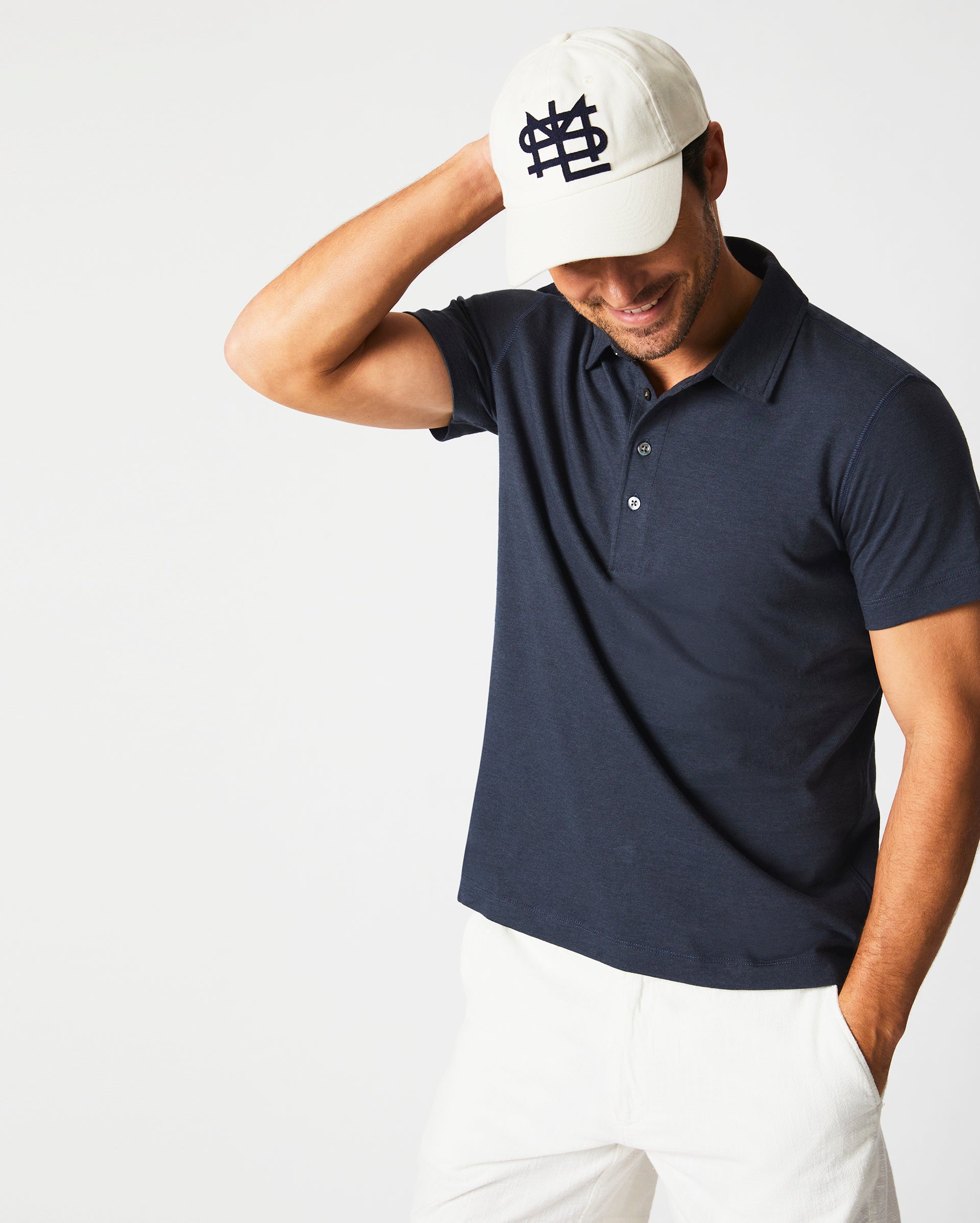 A smiling man in a navy polo shirt and white pants touches the brim of his white baseball cap, which has a dark logo on the front, while standing against a plain white background.