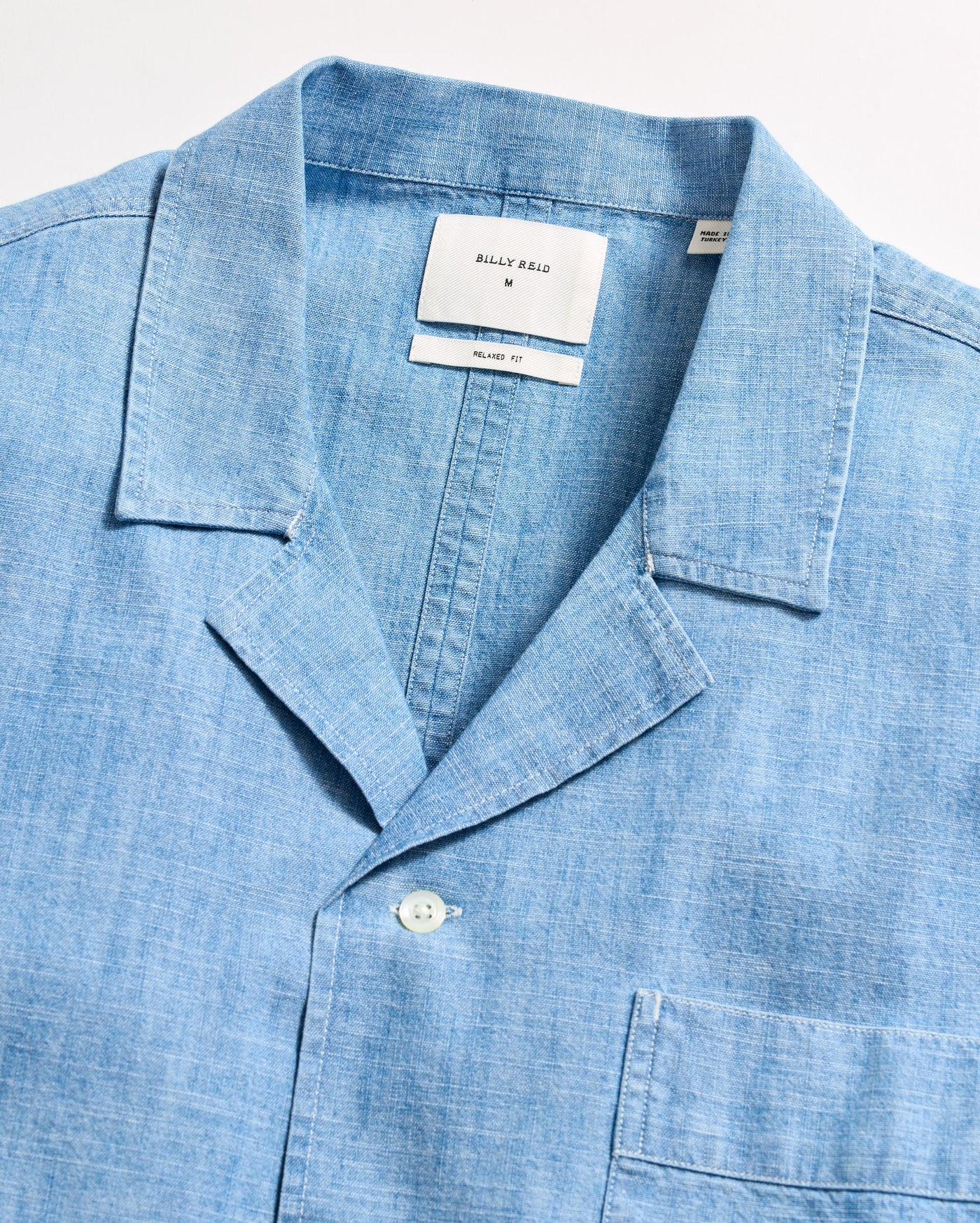 Light blue, short-sleeve button-up shirt made from a chambray or linen-like fabric, featuring a notched collar, single chest pocket, and visible white buttons. The shirt label reads Billy Reid.