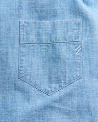 A close-up of a light blue denim shirt pocket with visible stitching and two small white embroidered lines near the upper right corner of the pocket.