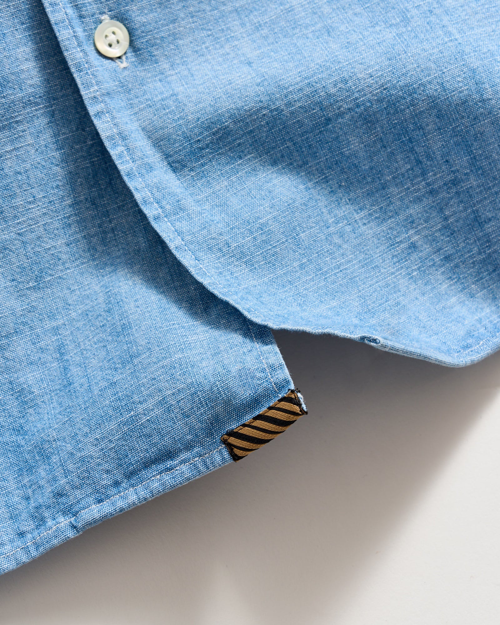 A close-up of the bottom hem of a light blue button-up shirt, showing a white button and a small brown and black striped fabric tag.