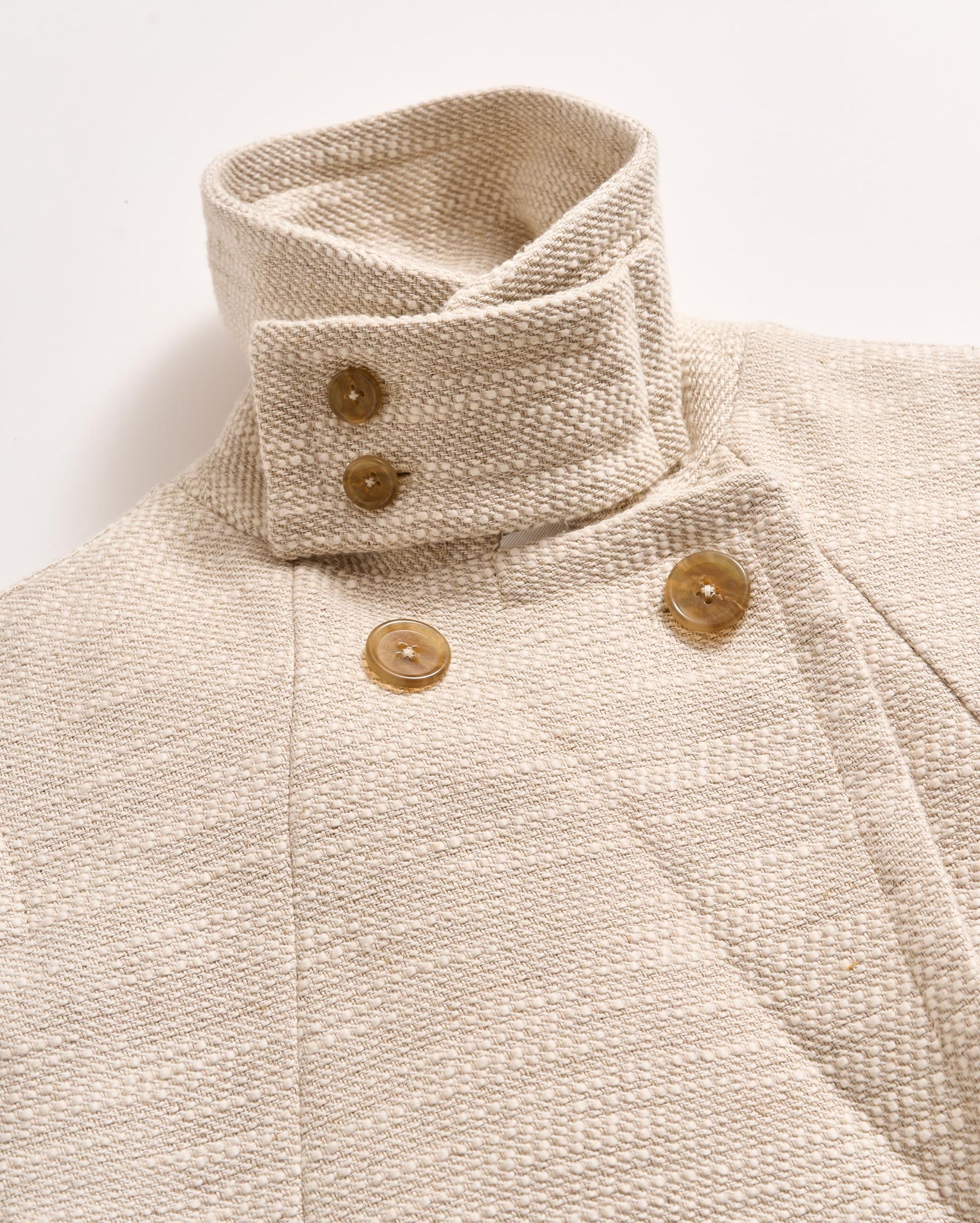 A close-up of a cream-colored textured coat with a high, structured collar and large tan buttons arranged in a double-breasted style against a plain white background.
