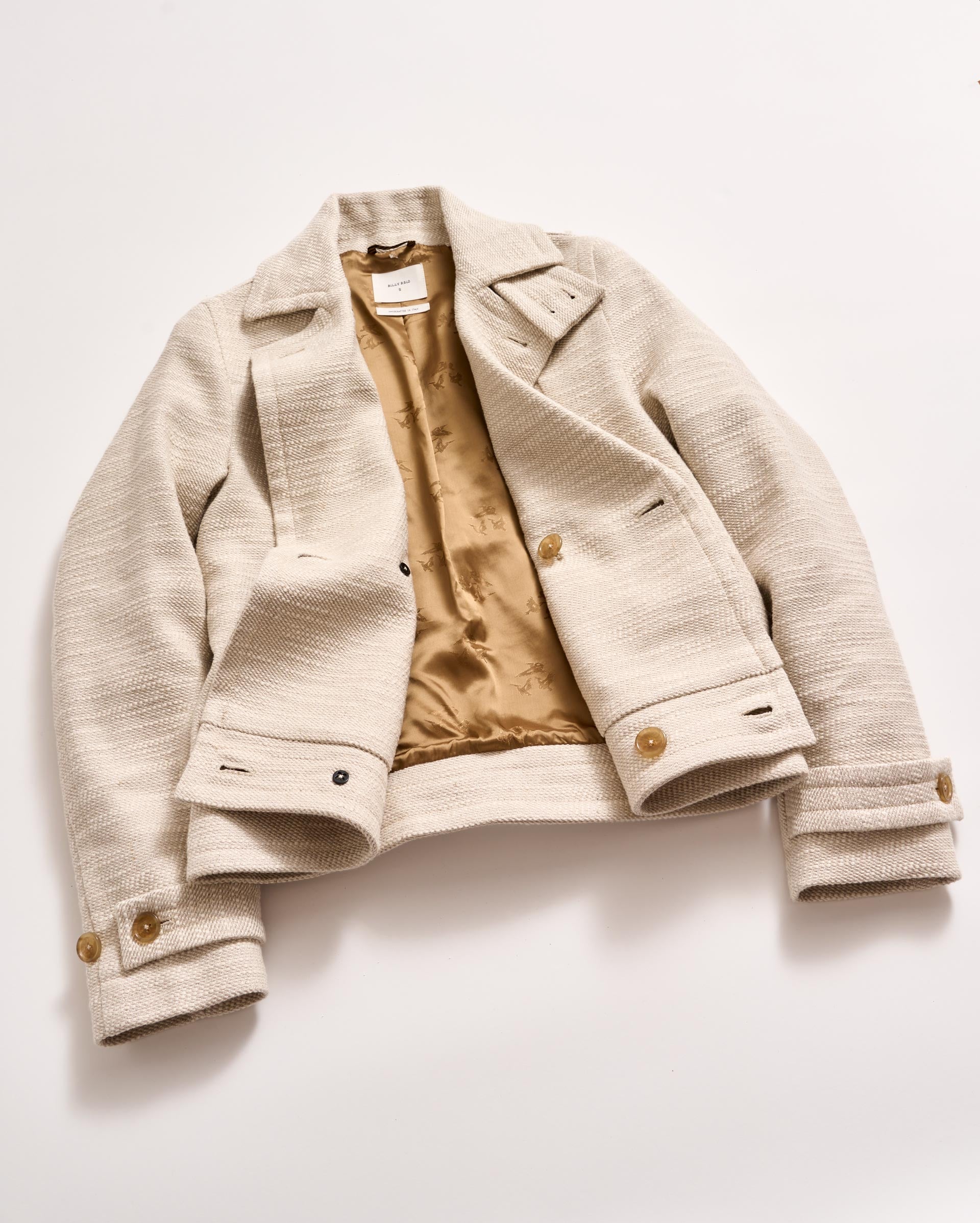 A beige textured women’s jacket with a tan satin lining, buttoned cuffs, lapel collar, and front pockets, laid flat on a white surface.