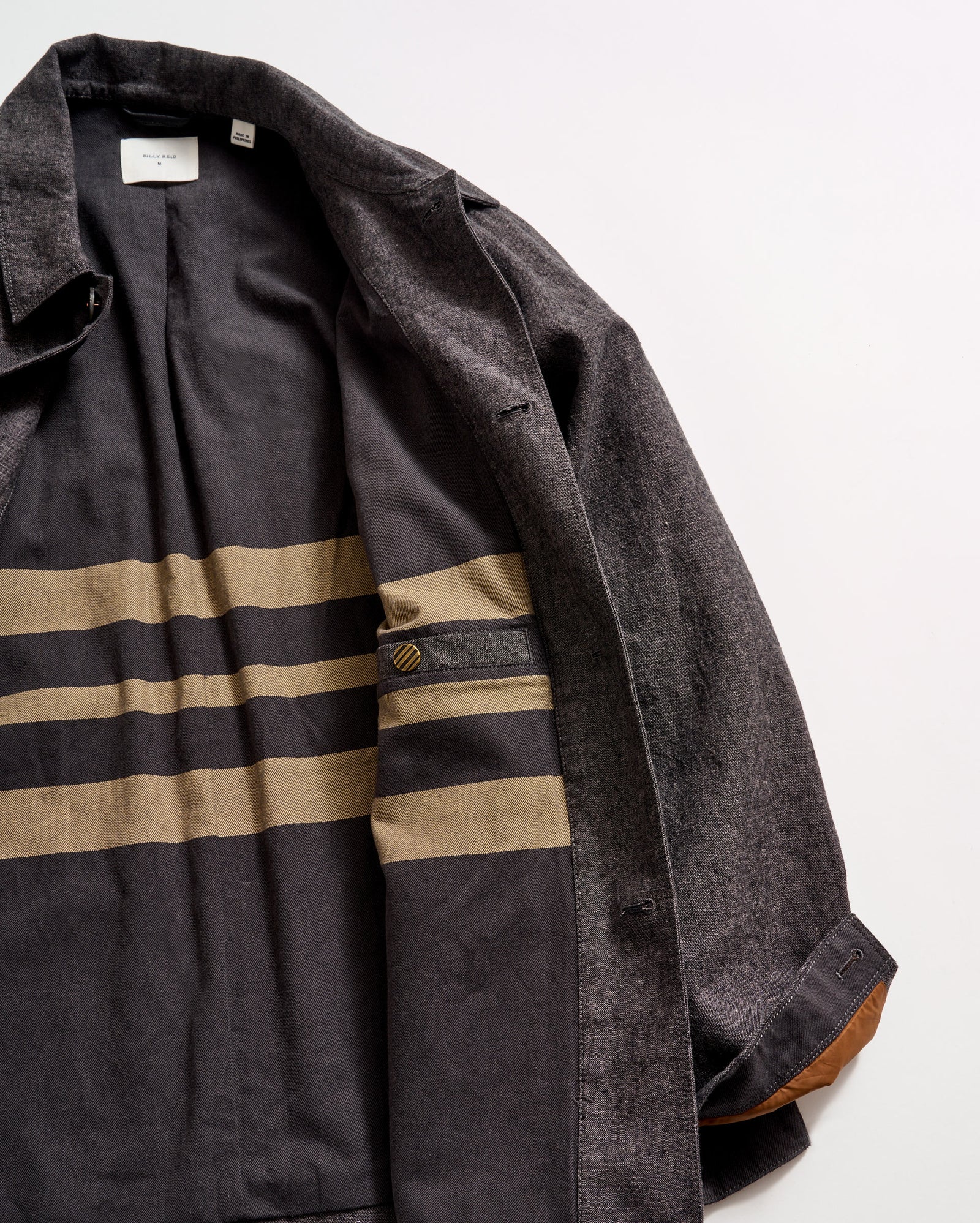 A dark gray coat is laid open, revealing its inner lining with two wide horizontal tan stripes and a small buttoned pocket. The coat has a brown accent on the inner cuff and is displayed on a light surface.
