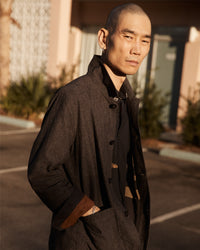 A man with a shaved head wearing a dark coat stands outdoors with his hands in his pockets, looking confidently at the camera. The background shows a building, doors, and some greenery in soft sunlight.