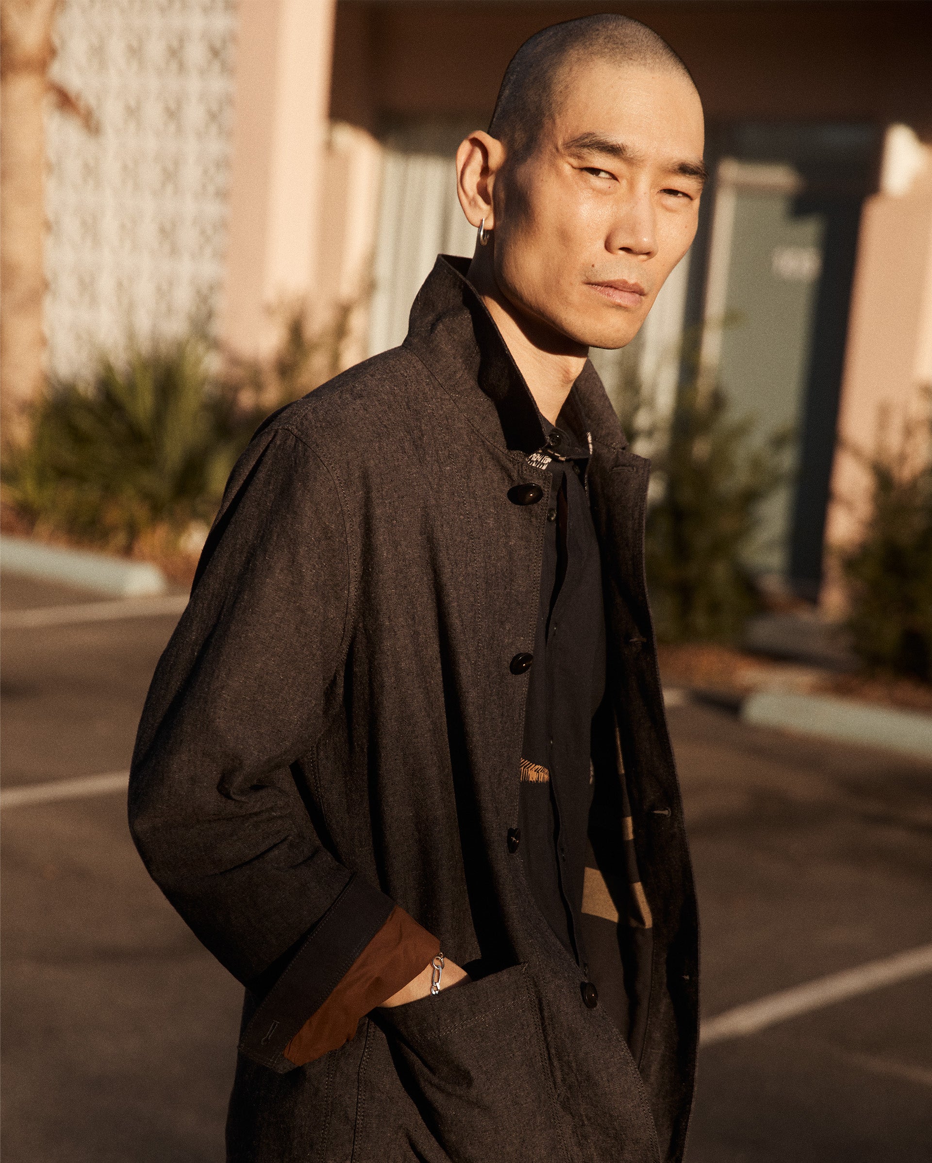 A man with a shaved head wearing a dark coat stands outdoors with his hands in his pockets, looking confidently at the camera. The background shows a building, doors, and some greenery in soft sunlight.