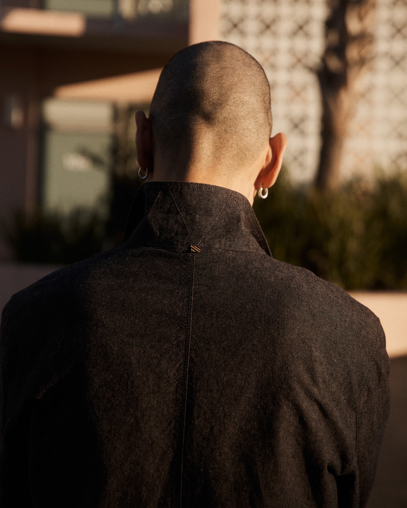 A person with a shaved head and hoop earrings is seen from behind, wearing a dark shirt. The background is softly blurred, showing a building, greenery, and warm sunlight.