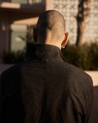 A person with a shaved head and hoop earrings is seen from behind, wearing a dark shirt. The background is softly blurred, showing a building, greenery, and warm sunlight.