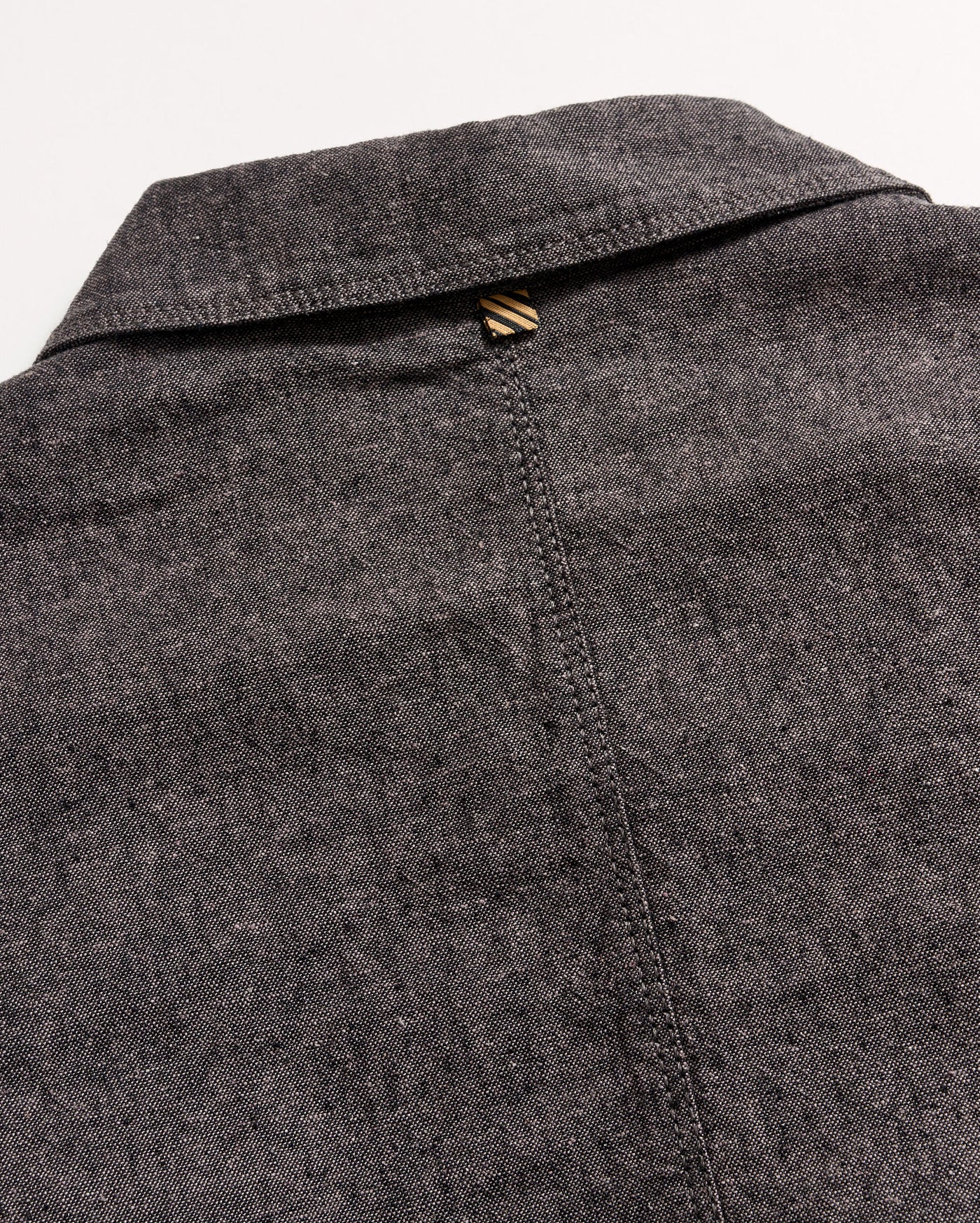 Close-up of the back of a grey textured collared shirt, showing a seam running down the center and a small brown and beige striped loop at the top below the collar.