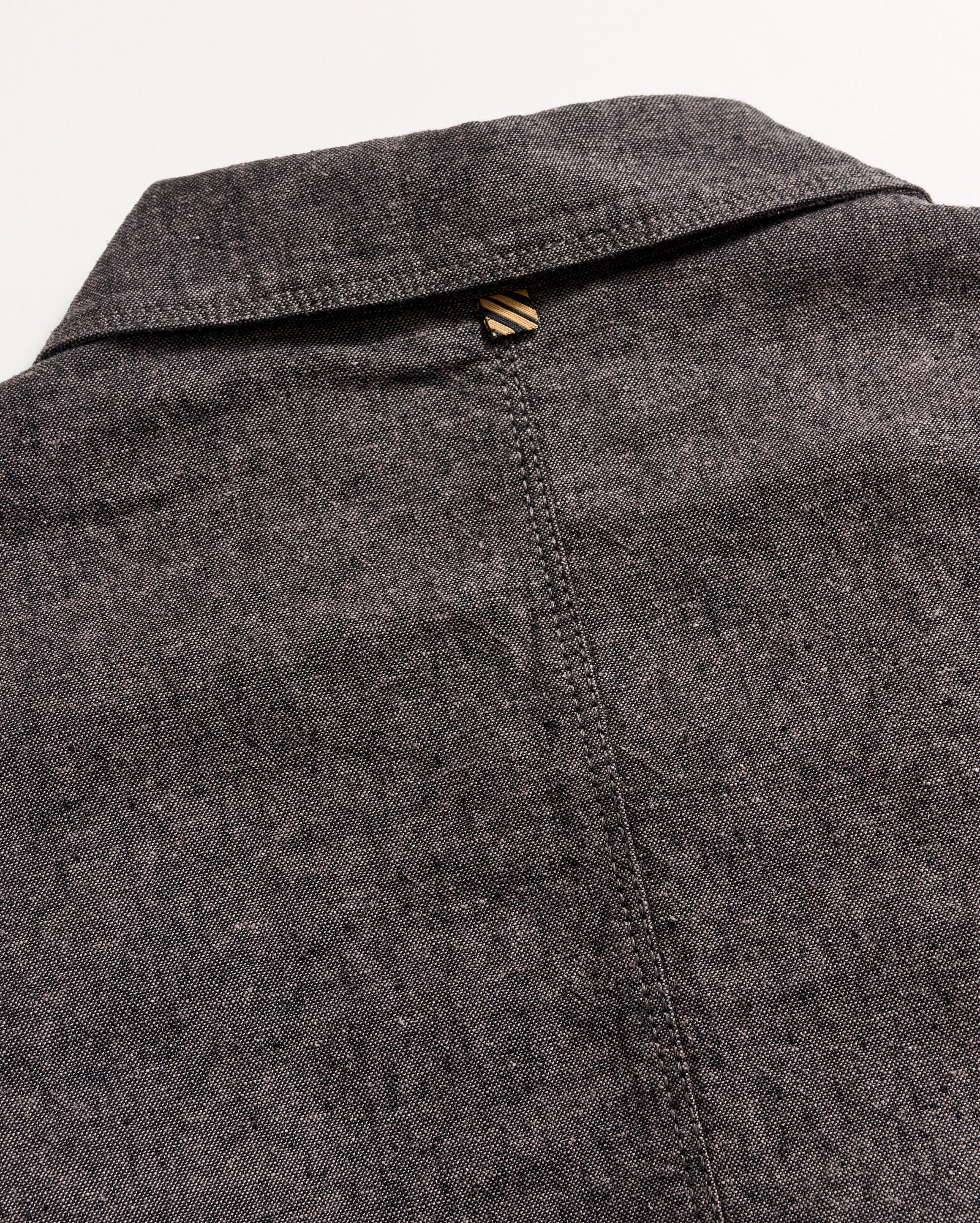 Close-up of the back of a grey textured collared shirt, showing a seam running down the center and a small brown and beige striped loop at the top below the collar.