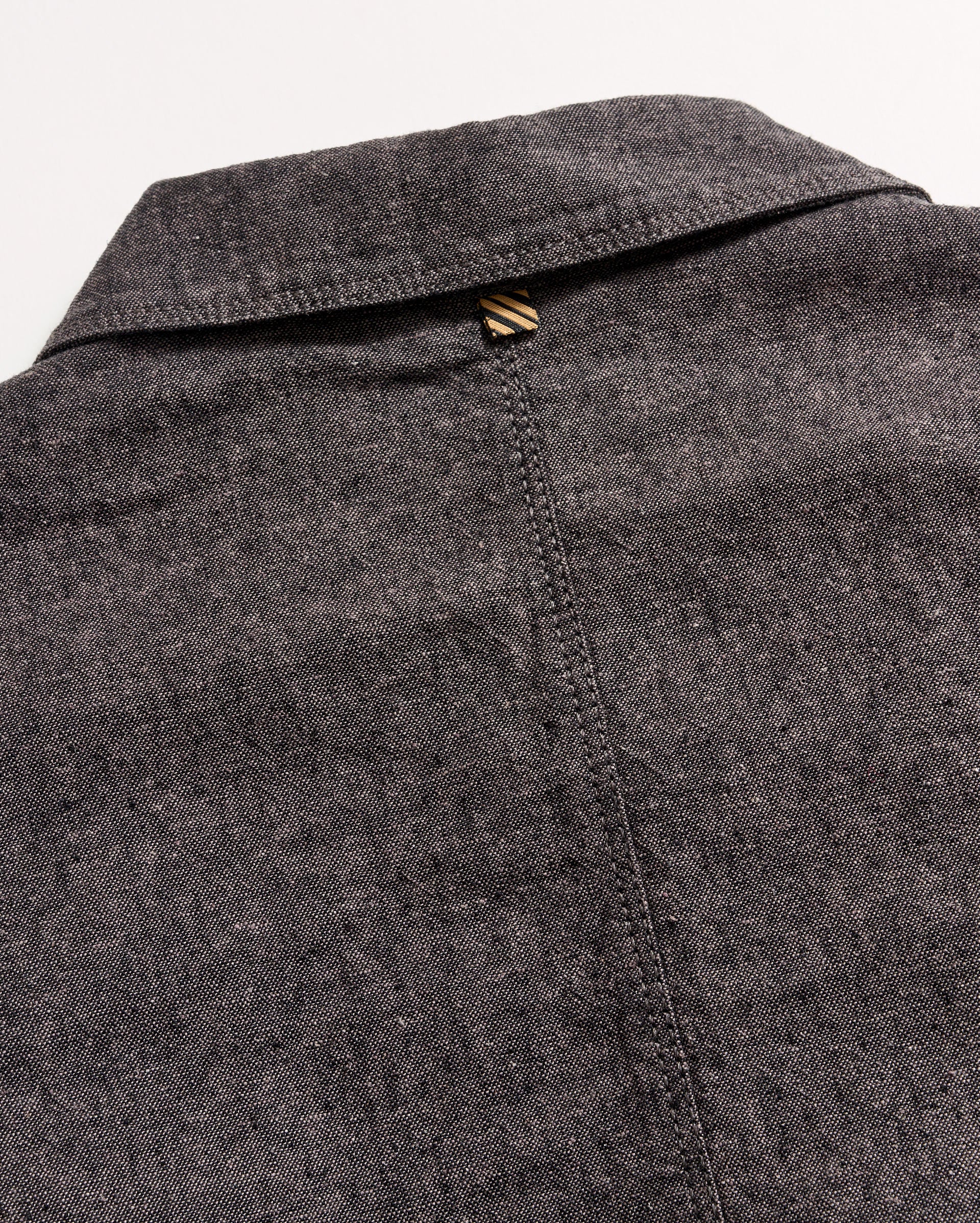 Close-up of the back of a grey textured collared shirt, showing a seam running down the center and a small brown and beige striped loop at the top below the collar.
