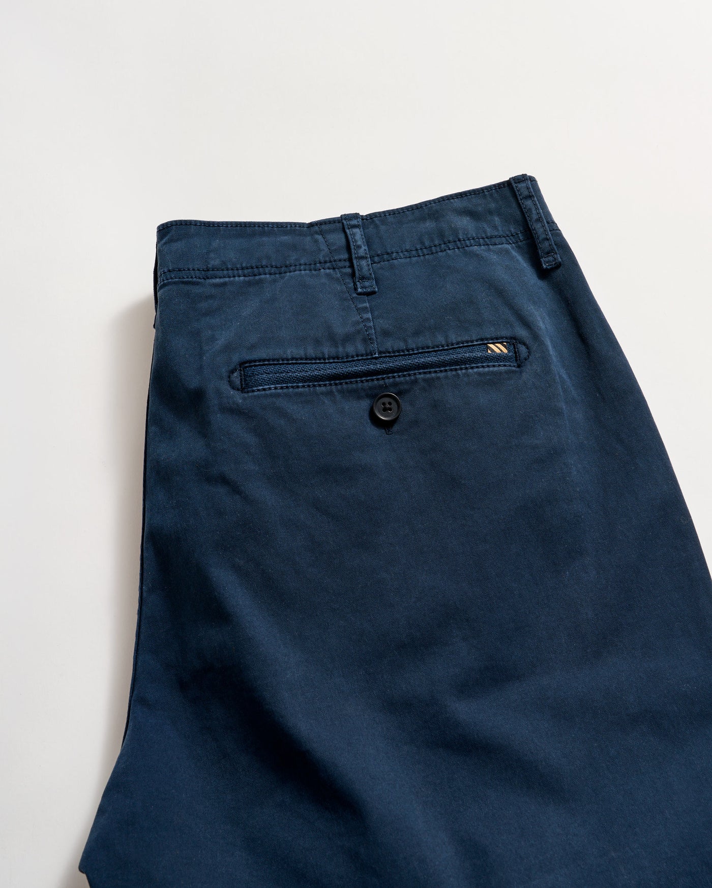A pair of dark blue pants featuring a back welt pocket with a button closure, a small stitched detail near the pocket, and a visible belt loop, laid flat on a light background.