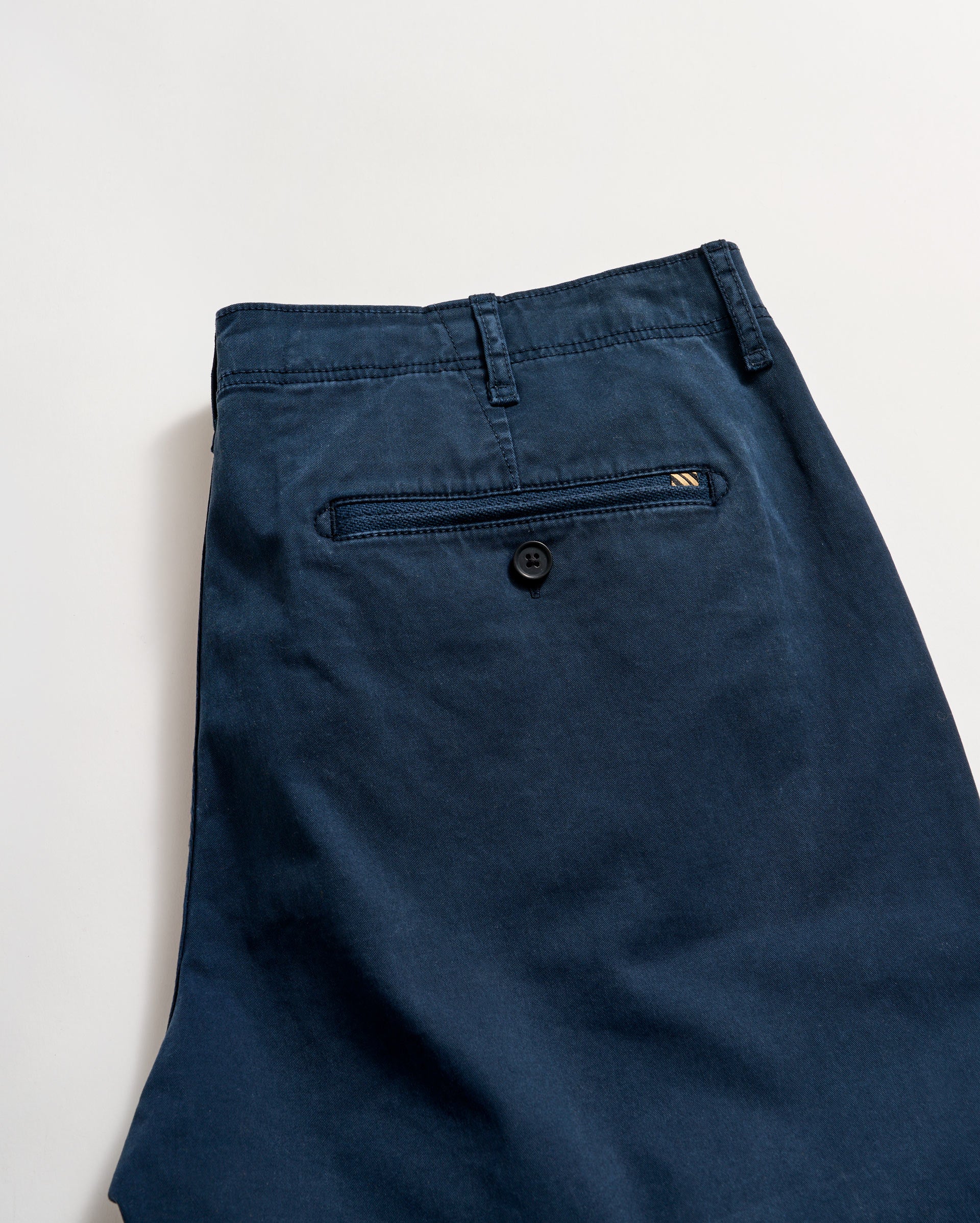 A pair of dark blue pants featuring a back welt pocket with a button closure, a small stitched detail near the pocket, and a visible belt loop, laid flat on a light background.