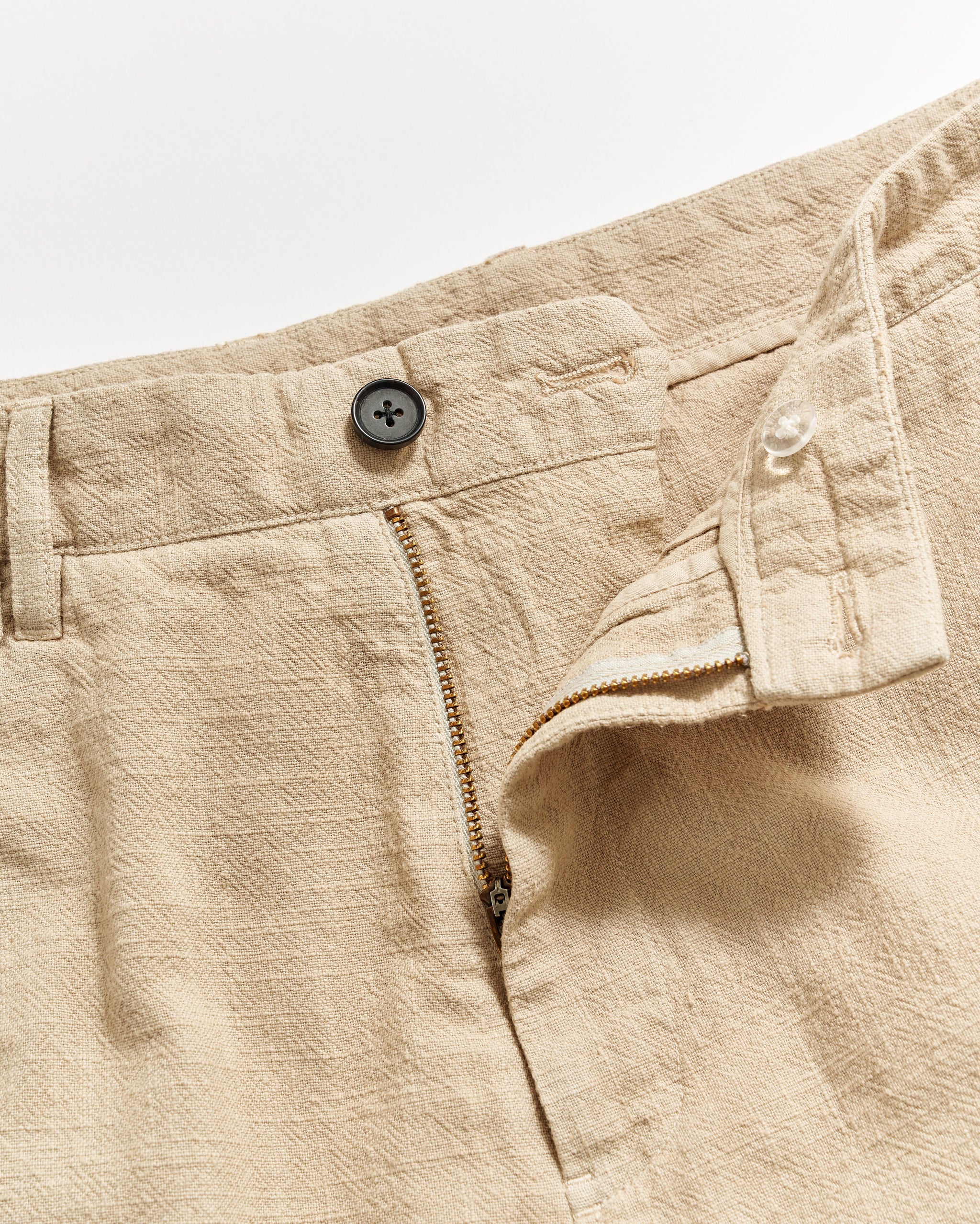 Beige textured fabric pants with a partially open zipper fly, a black button at the waistband, and an inner button closure, shown on a white background.