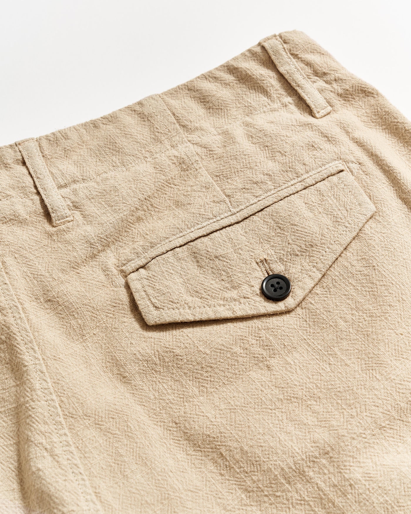 Close-up of beige textured pants showing a back pocket with a flap and a black button, along with belt loops and visible stitching details.