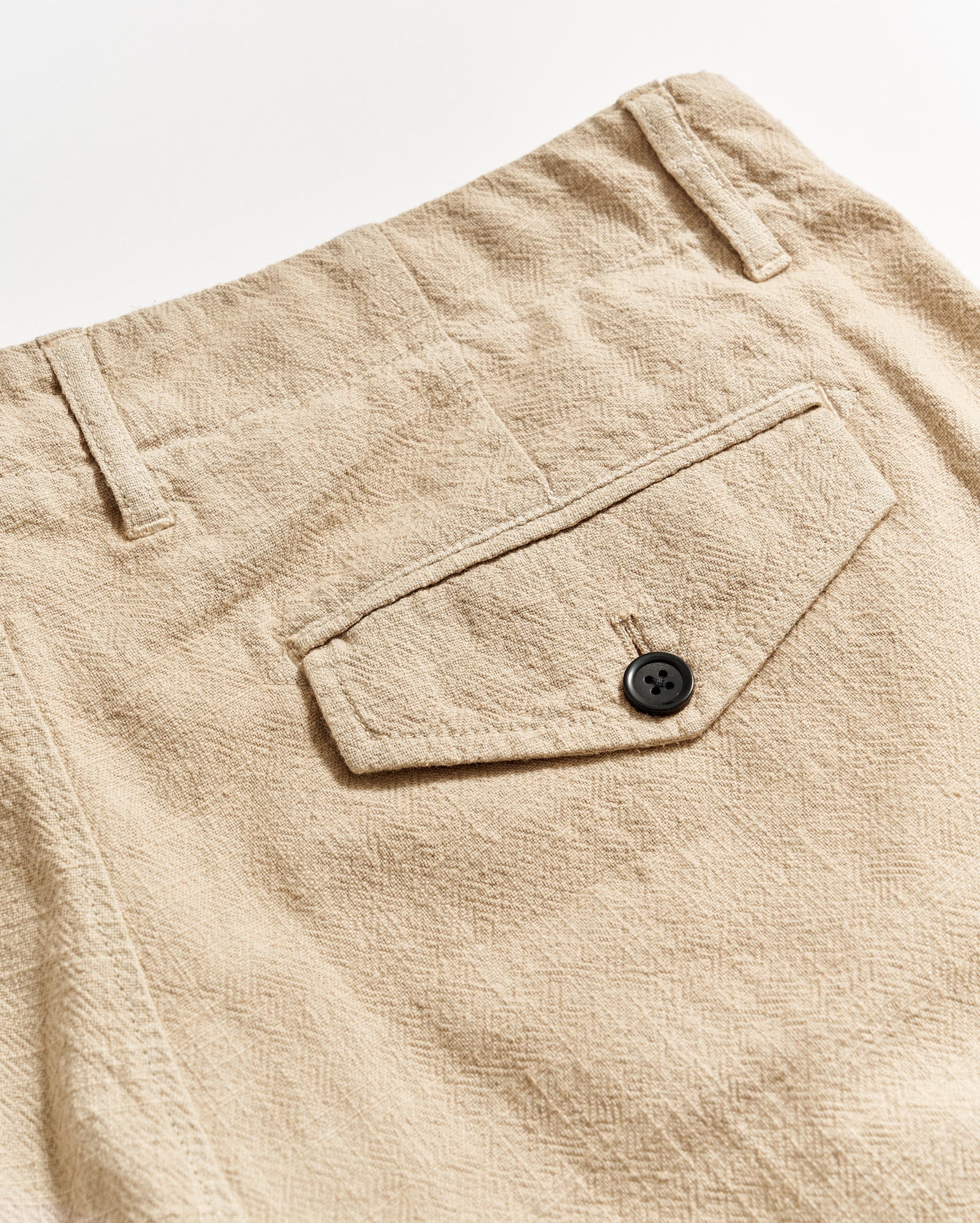 Close-up of beige textured pants showing a back pocket with a flap and a black button, along with belt loops and visible stitching details.