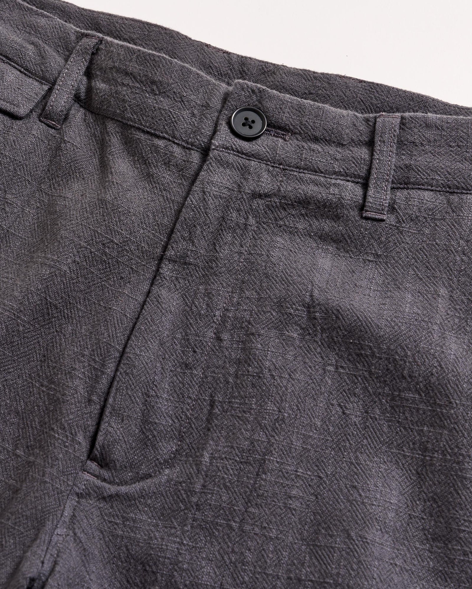 Close-up of the waistband and upper section of a pair of gray textured pants with a button closure and belt loops, set against a light background.