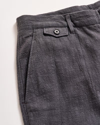 Close-up of the upper section of a pair of textured gray pants, featuring a waistband with a belt loop, a small decorative flap with a black button, and a pleated design near the pocket.