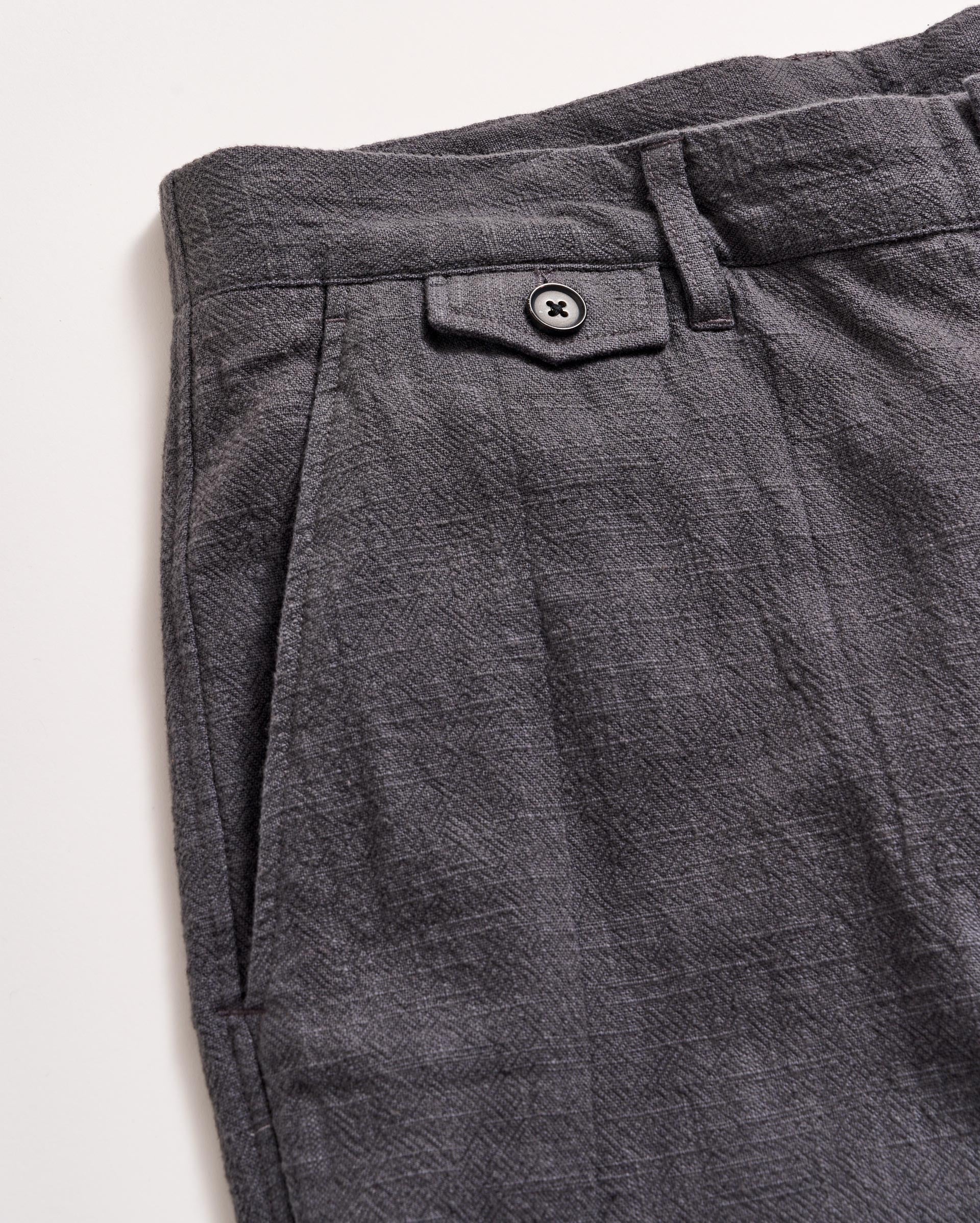 Close-up of the upper section of a pair of textured gray pants, featuring a waistband with a belt loop, a small decorative flap with a black button, and a pleated design near the pocket.