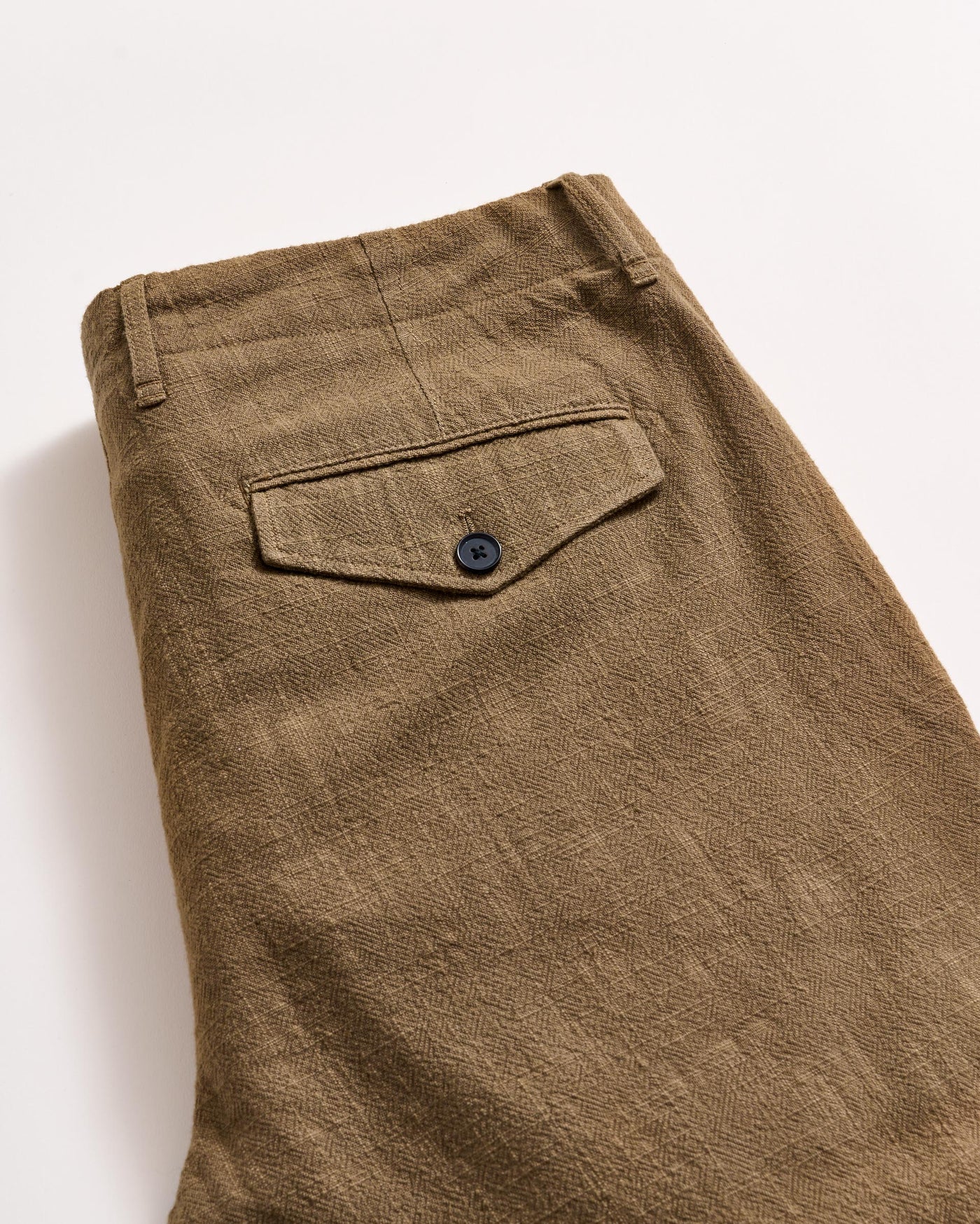 A close-up of a pair of folded brown textured pants, showing a back pocket with a black button and a belt loop on a light background.