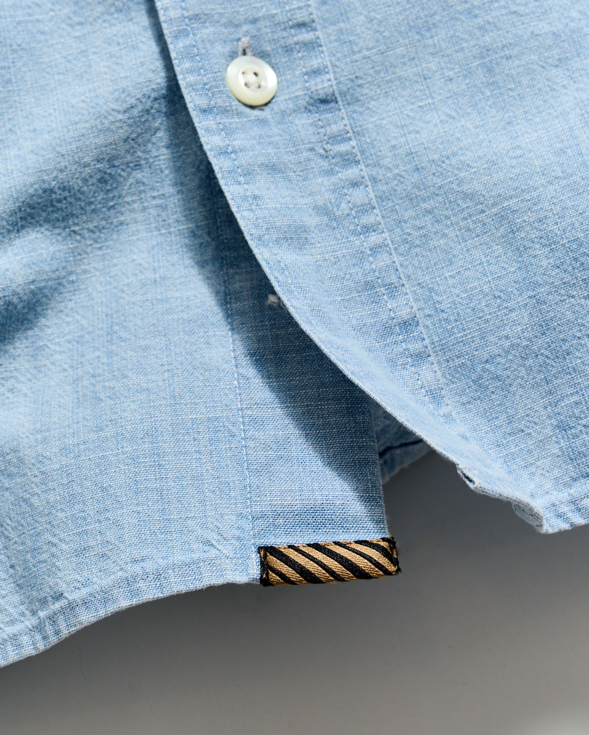Close-up of a light blue denim shirt with a button and brand label on a gray background