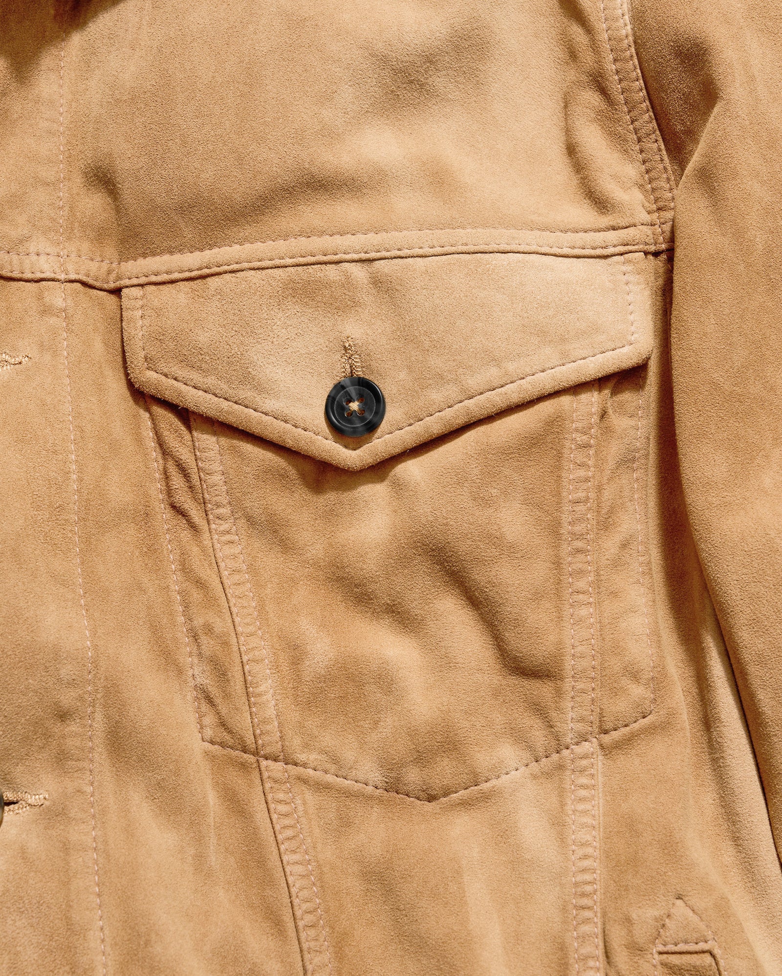 Close-up of a tan suede jacket with a flap pocket and a dark button, showing the texture and stitching details.