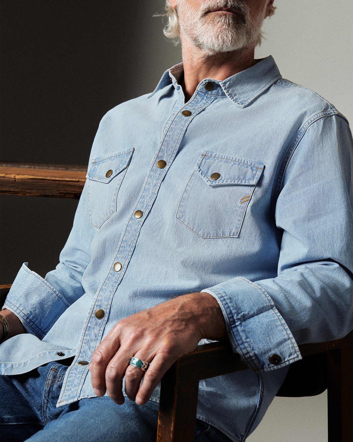 Man wearing a light blue denim shirt sitting on a wooden chair.