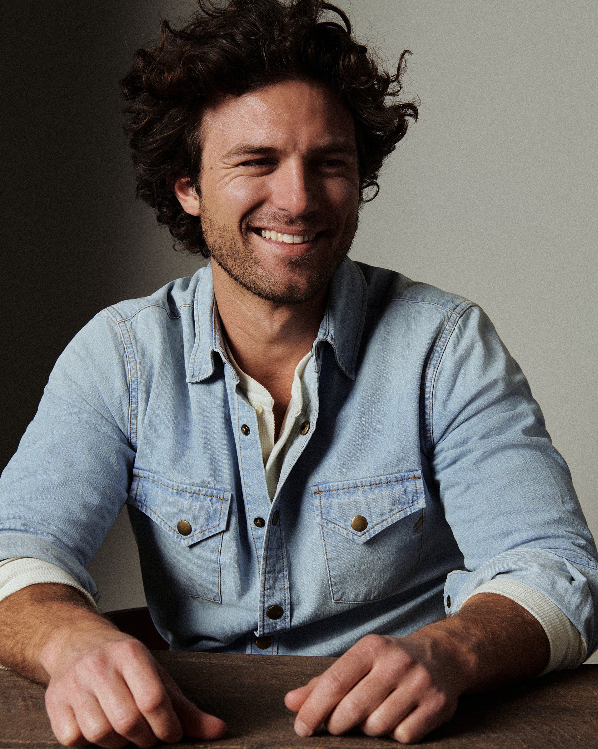 Man wearing a light blue denim shirt sitting at a table with a neutral background
