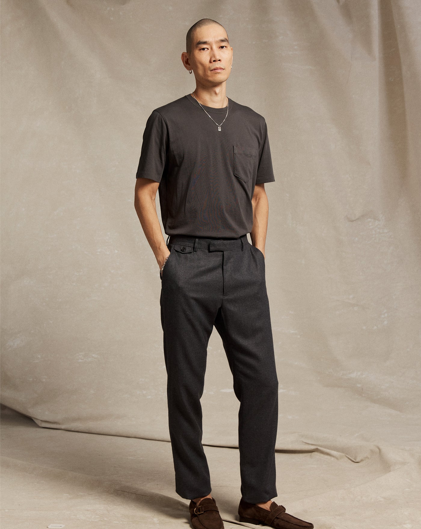 A person with a shaved head stands against a beige fabric backdrop, wearing a dark short-sleeve t-shirt, dark tailored pants, brown shoes, and a necklace, with hands in their pockets and a neutral expression.