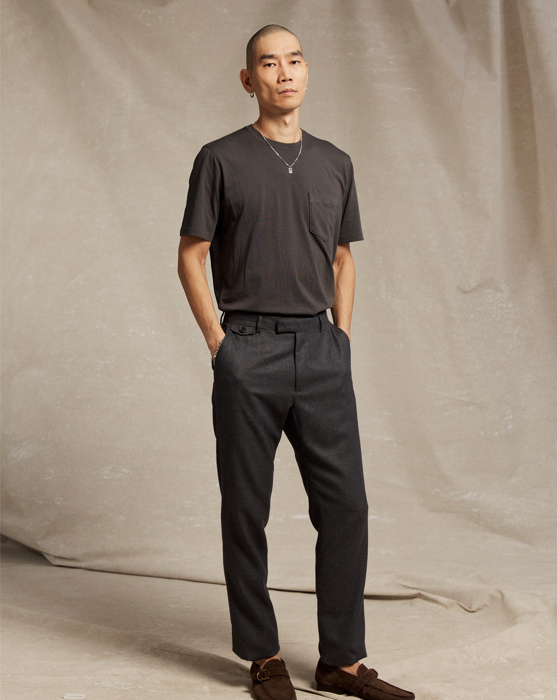 A person with a shaved head stands against a beige fabric backdrop, wearing a dark short-sleeve t-shirt, dark tailored pants, brown shoes, and a necklace, with hands in their pockets and a neutral expression.