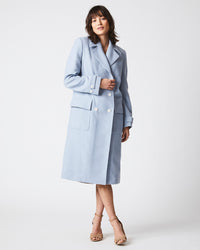 A woman stands against a plain white background wearing a light blue, knee-length coat with large buttons and front pockets. She is also wearing tan high-heeled sandals.